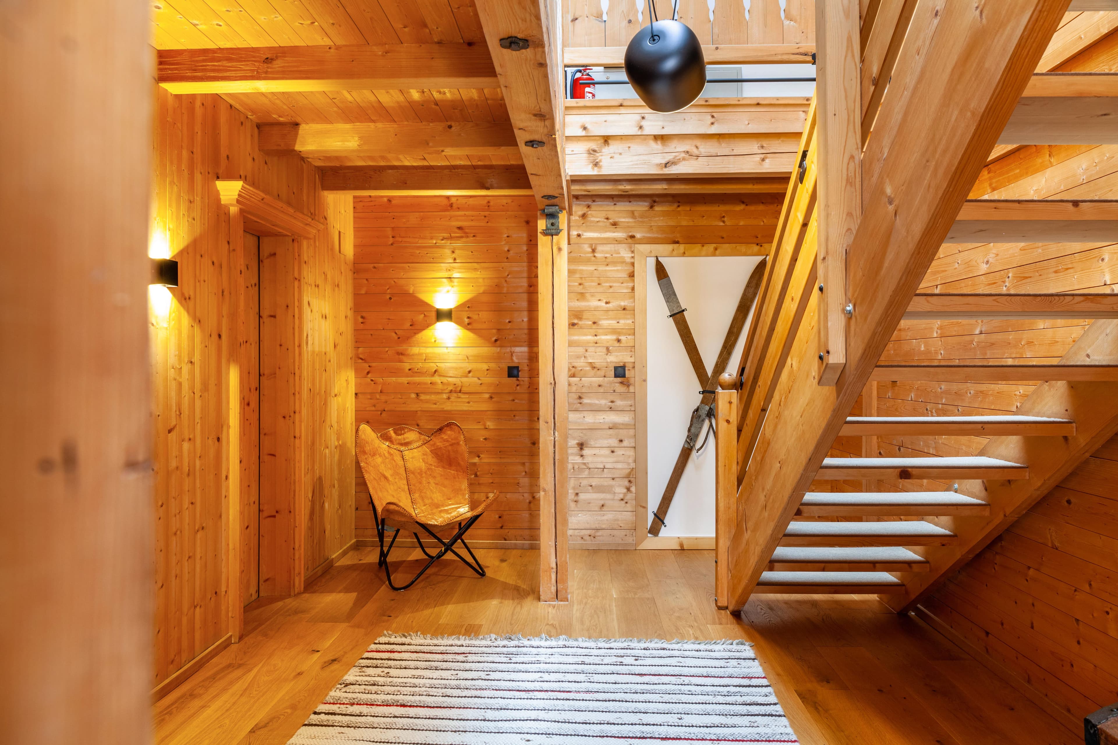 Gemütliches Interieur aus Holz mit einer Treppe, einem Schmetterlingsstuhl und einem gestreiften Teppich. Die warme Beleuchtung unterstreicht das rustikale Ambiente.