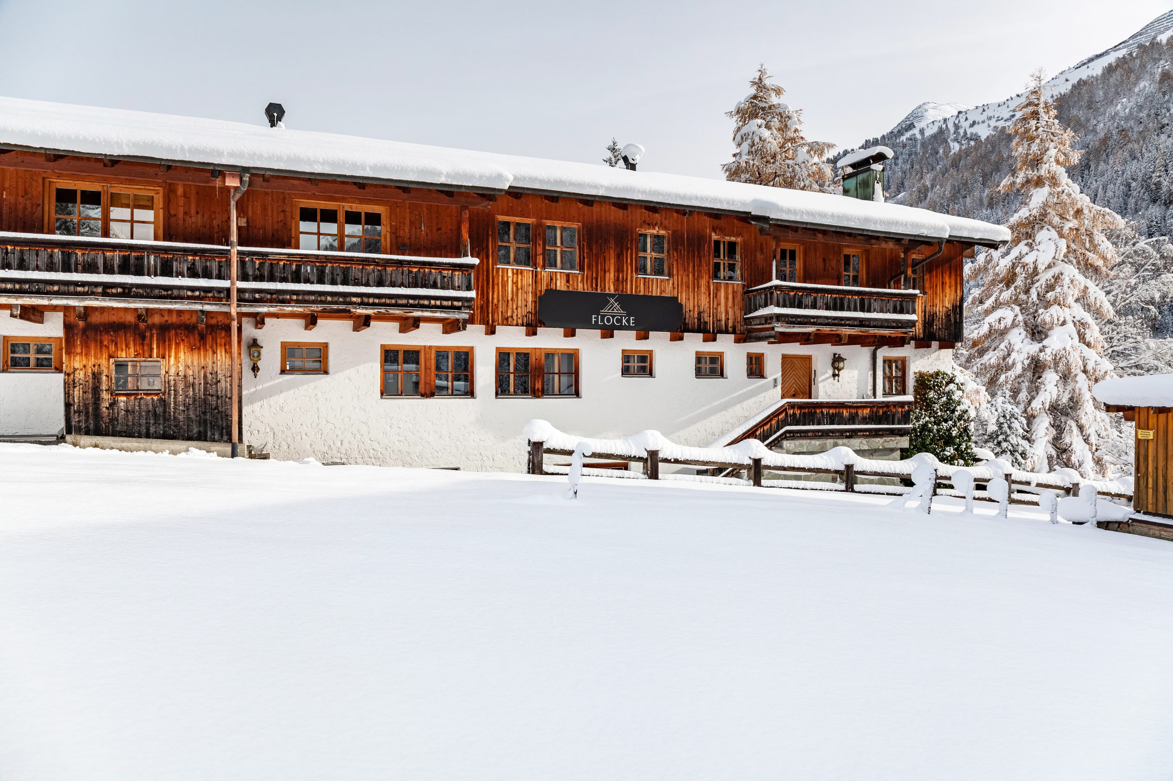 Ein verschneites Holzchalet mit Balkonen, umgeben von schneebedeckten Bäumen und Bergen unter einem klaren Himmel.