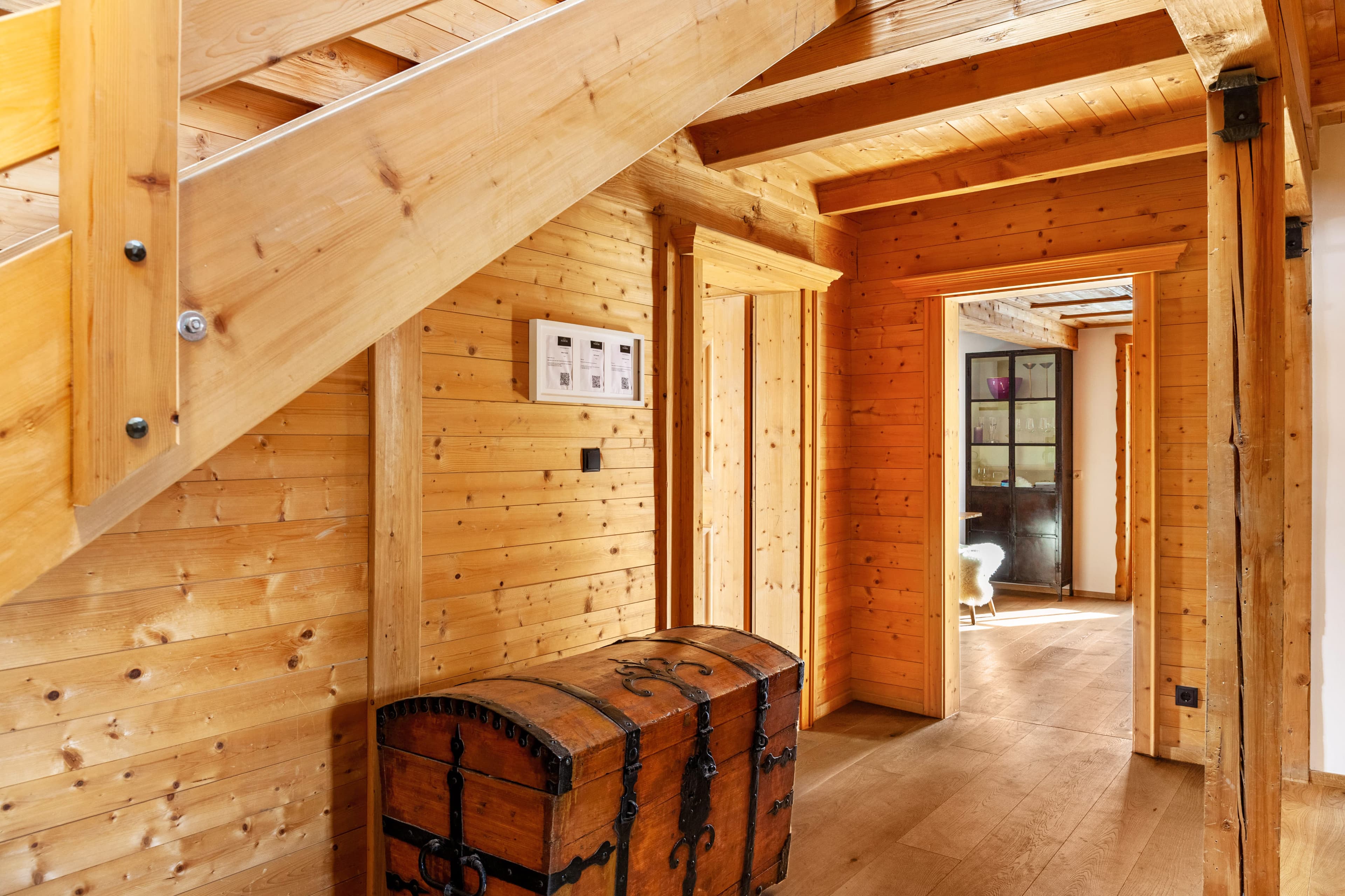 Holzflur mit sichtbaren Balken, einer alten Truhe und gerahmten Bildern an der Wand. Eine Tür führt zu einem Zimmer mit einem Fenster und einem Stuhl.