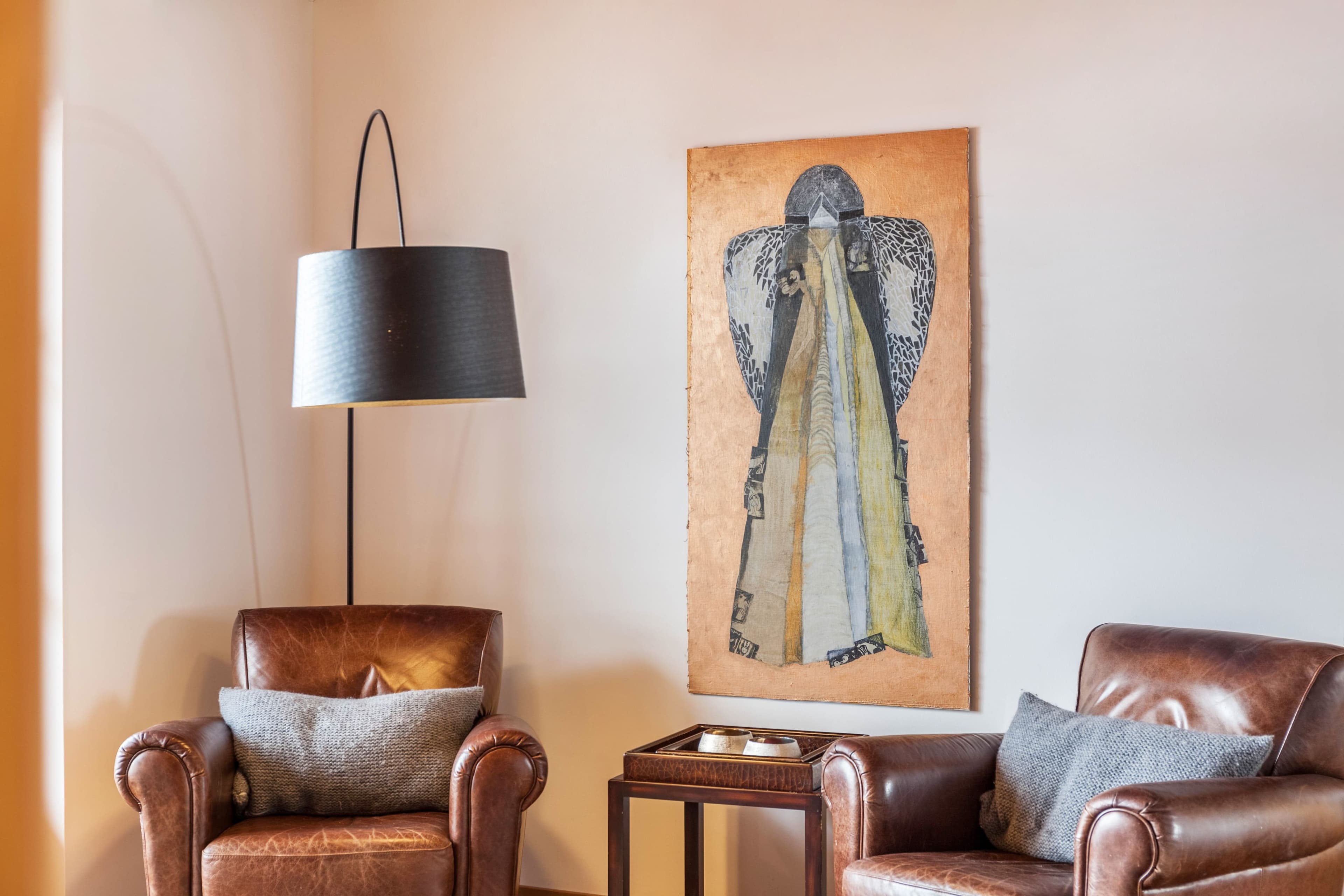 Gemütliches Zimmer mit zwei braunen Ledersesseln, einer Stehlampe und einem Wandgemälde, das abstrakte Formen darstellt. Auf einem kleinen Tisch stehen Bücher.